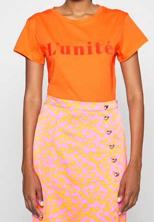 Femme portant un t-shirt orange vif avec le texte rouge "L'unité", assorti d'une jupe rose ornée de motifs de cœurs jaunes et de boutons en forme de cœur.