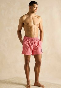 Ružové plavky vyrobené z ľahkého materiálu s elastickým pásom, bočnými vreckami a malým modrým emblémom na ľavej nohe.