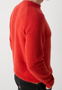 Roter Strickpullover mit rundem Ausschnitt und gerippten Bündchen, mit weicher Textur und figurbetontem Schnitt, getragen zu schwarzen Jeans.