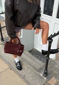 Borsa a tracolla bordeaux intrecciata con doppio manico, mocassini neri lucidi, calzini bianchi e una giacca di pelle marrone; soggetto in posa su gradini di pietra.