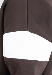 Haut à capuche marron avec une bande blanche horizontale. Fabriqué en tissu doux avec une texture lisse et une coupe décontractée, idéal pour un usage quotidien.