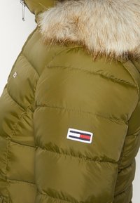 Olivově zelená péřová bunda s límcem z umělé kožešiny, prošívanou texturou a malou vyšitou nášivkou v červené, bílé a modré barvě na rukávu.