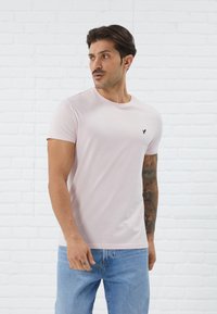 Camiseta de algodón rosa claro con cuello redondo, mangas cortas y un pequeño logo negro en la parte superior izquierda. Llevada con jeans azul claro.