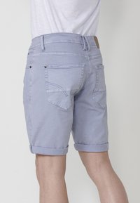 Aizmugurējais skats uz personu, kura valkā gaiši zilos sarullētos džinsus un baltu kreklu, stāvot pret vieglu fonu.