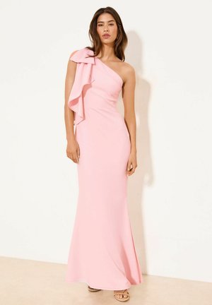 Vrouw in een lichtroze, een-schouder lange jurk met een grote volant op de schouder, staand tegen een effen witte muur.