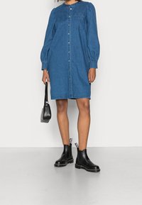 Blå denimklänning med långa puffärmar, knappstängning, bröstficka och avslappnad passform, kombinerad med svarta Chelsea-stövlar och en liten väska.