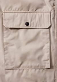 Abrigo beige con un gran bolsillo de solapa. La tela suave y texturizada cuenta con un cierre de botón y detalles de costura en contraste.