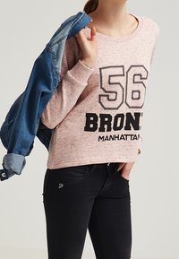 Felpa rosa chiaro con testo stampato in nero "56 BRONX MANHATTAN". Abbinata a una giacca di jeans, indossata sopra jeans neri. Texture morbida.