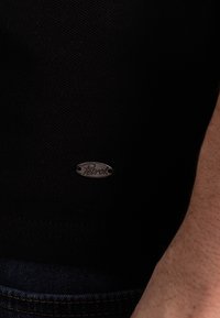 Černé texturované tričko s krátkým rukávem, které má malé stříbrné oválné logo se slovem "Petrol" blízko lemu, spárované s modrými džínami.