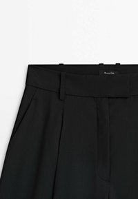 Sorte bukser med en bred linning, der har sidelommer med folder og en glat tekstur. Et mærke er synligt på linningen.