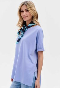 Jasnofioletowa luźna bluzka z krótkimi zwiniętymi rękawami i bocznymi rozcięciami, w zestawieniu z czarno-niebieskim wzorzystym szalem. Gładka, miękka tekstura.