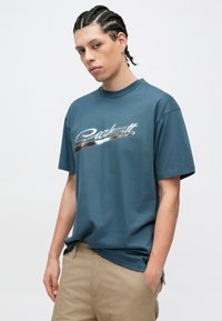 Camiseta de manga corta color verde azulado oscuro con un logotipo plateado brillante de "Carhartt" en el pecho y un ajuste relajado. Material de algodón suave.