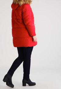 Červený prošívaný kabát s kapucí lemovanou kožešinou, kapsami na zip, prošívaným designem a volným střihem; kombinovaný s tmavými džínami a černými botami na podpatku.
