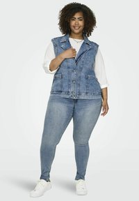 Chaleco de mezclilla en azul claro con cuello, botones en el frente y dos bolsillos delanteros, combinado con pantalones ajustados de mezclilla azul claro y zapatillas blancas.