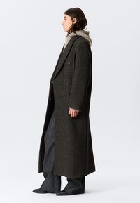 Dark green, long wool coat with a double-breasted design, large lapels, and textured fabric. Worn over a gray hoodie and paired with wide-leg pants.