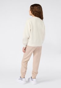Niño con pelo rizado y marrón de espaldas, vistiendo un sudadera crema, pantalones deportivos beige y zapatillas blancas, sobre un fondo blanco.