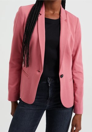 Kvinde iført en pink blazer over en sort top med mørkeblå jeans, stående foran en ensfarvet lys baggrund.