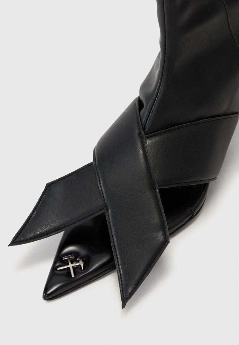 Svart läderankelboot med spetsig tå, korskorsad design och metallisk logotypaccent fram. Slät textur genom hela skon.