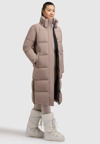 Langer Daunenmantel in hellem Beige mit hohem Kragen, Druckknöpfen, Fronttaschen und kontrastierenden schwarzen Reißverschlussakzenten. Kombiniert mit weißen Stiefeln.