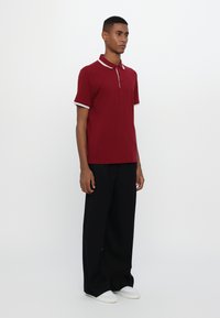 Burgunderfarget polo skjorte med hvite detaljer, med skjortekrage og tre knapper, kombinert med vide svarte bukser og hvite sneakers.