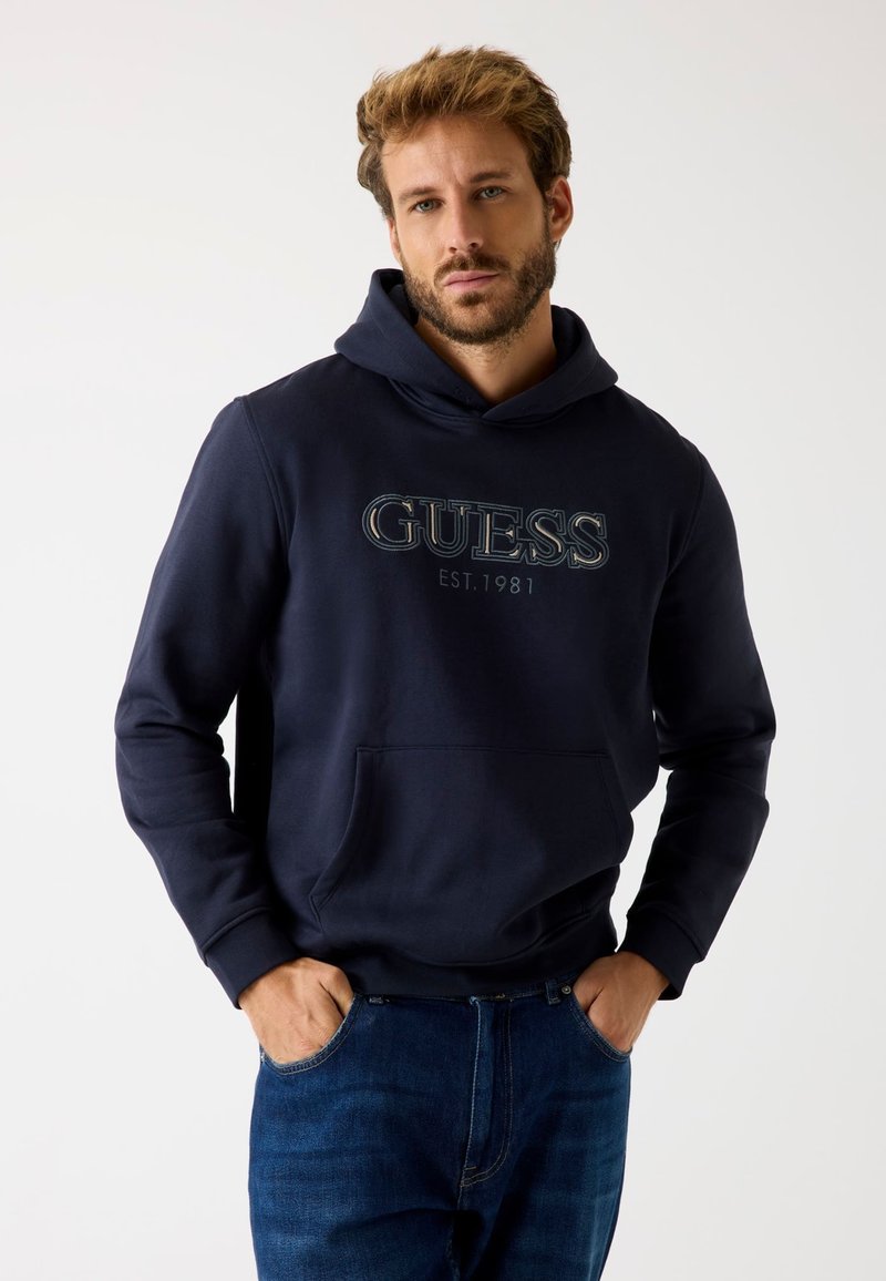 Tengerészkék kapucnis pulóver elülső zsebből és "GUESS EST. 1981" logóból. Pamut anyag, normál fazon, bordás mandzsetta és szegély.