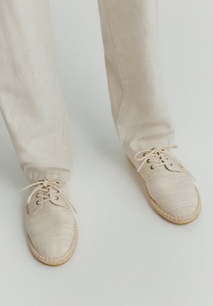 Scarpe in tela beige con lacci indossate con pantaloni beige chiaro abbinati su uno sfondo neutro.