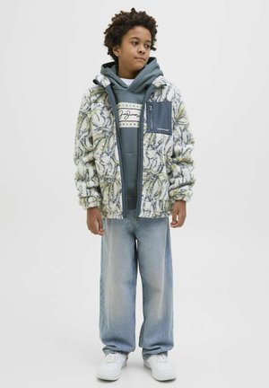 Veste blanche duveteuse avec un motif floral jaune et vert, superposée à un sweatshirt gris, accompagnée de jeans larges bleu clair et de baskets blanches.