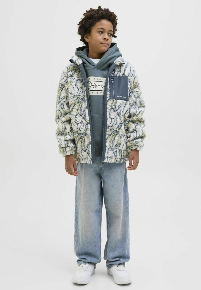 Chaqueta blanca esponjosa con un patrón floral amarillo y verde, superpuesta a una sudadera gris, combinada con unos pantalones vaqueros de pierna ancha de color azul claro y zapatillas blancas.