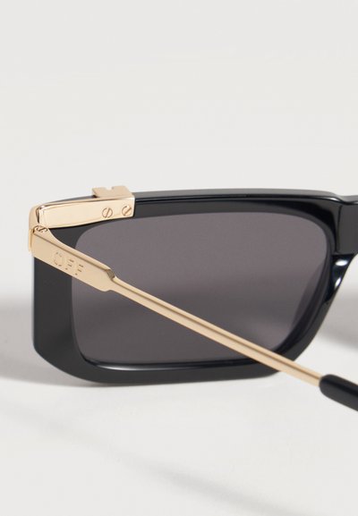 Gros plan de lunettes de soleil noires avec des verres rectangulaires foncés et des branches métalliques dorées gravées "OFF."