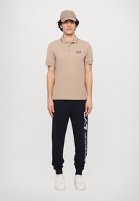 Tan polo shirt with black trim, logo on chest; navy sweatpants with white logo and text; white sneakers; coordinating tan cap.