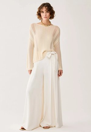 Donna indossa un maglione lavorato a maglia color crema, pantaloni bianchi a gamba larga con fiocco laterale, collana di perline dorate e sandali, in piedi davanti a uno sfondo chiaro.