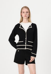Black cardigan with cream trim, featuring a shawl collar, front pockets, and button closure. Worn over a white top and black shorts.