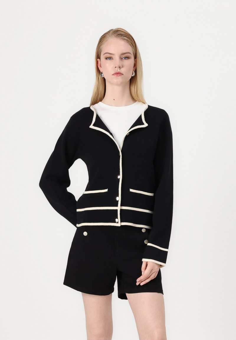 Cardigan nero con rifiniture crema, caratterizzato da un colletto a scialle, tasche frontali e chiusura a bottoni. Indossato sopra una maglietta bianca e pantaloni corti neri.