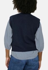 Marineblauer gestrickter Weste mit kurzen Ärmeln über einem gestreiften Hemd. Die Weste hat eine robuste Textur und eine taillierte Passform, kombiniert mit Jeans.