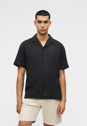 Mann trägt ein schwarzes Kurzarm-Hemd mit Knöpfen und beige knielange Shorts vor einem schlichten hellen Hintergrund.