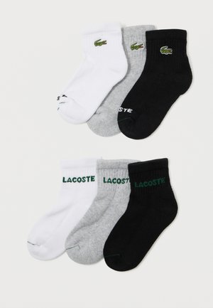 Šest párů bílých, šedých a černých ankletových ponožek Lacoste s vyšitými zelenými krokodýlími logy a textem "LACOSTE" na manžetách.