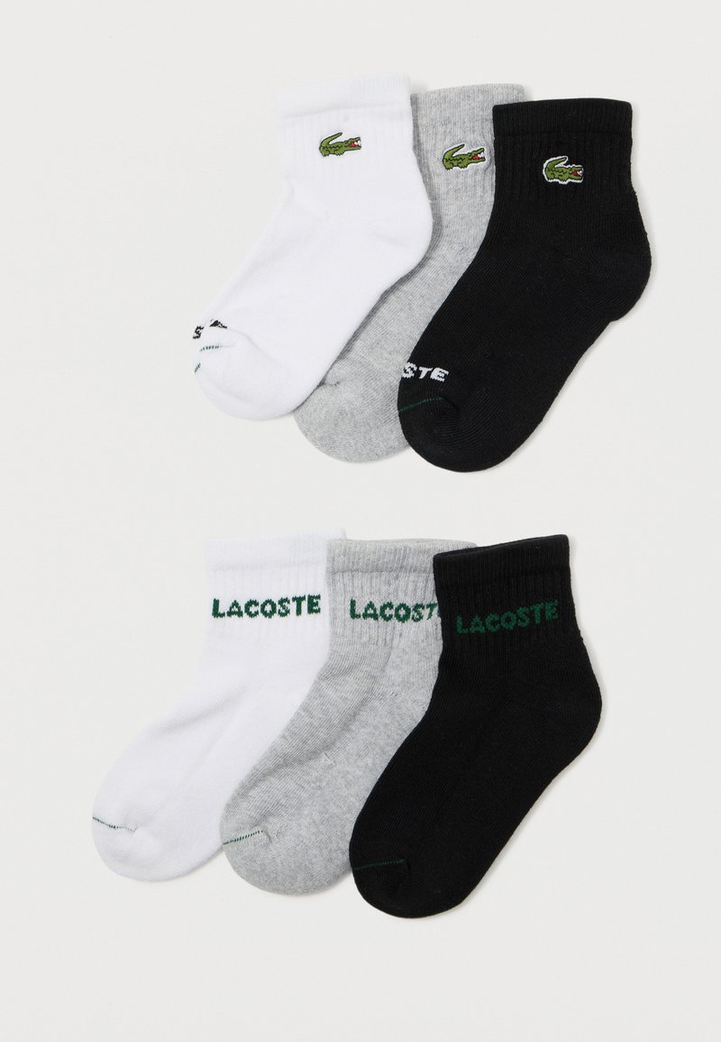 Seis pares de meias de tornozelo brancas, cinzentas e pretas da Lacoste com logos de crocodilo bordados em verde e o texto "LACOSTE" nos punhos.