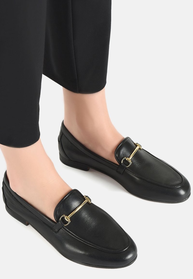 Pieds portant des mocassins en cuir noir avec un détail de barre métallique dorée, associés à un pantalon court noir sur fond blanc.