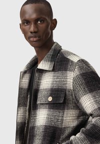 Veste à carreaux noir et blanc en mélange de laine avec un col évasé, fermeture éclair à l'avant et poche poitrine avec un détail de bouton.