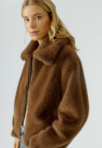 Blonde vrouw in een bruine donzige ritsjack met een grote kraag en blauwe spijkerbroek, die iets opzij kijkt tegen een effen achtergrond.