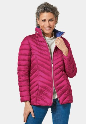 Fuchsia pufferjack met chevronpatroon, hoge kraag, ritssluiting en contrasterende blauwe binnenkant. Model draagt deze met een casual top en jeans.