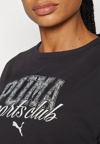 Musta puuvillapaita, jossa on pyöreä pääntie ja suuri "PUMA sportsclub" -grafiikka kuluneen metallinhohtoisen hopean ja valkoisen värissä.