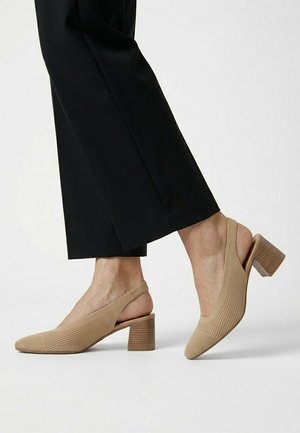 Zapatos beige con tacón bajo de bloque y tira en el talón, usados con pantalones negros cortos sobre fondo claro.
