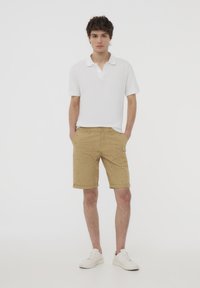 Polo bianco, pantaloni beige con piccoli motivi, e sneakers bianche. Il tessuto appare liscio; i pantaloni presentano risvolti e tasche frontali.