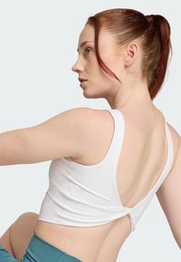 Top deportivo blanco sin mangas con diseño de torsión en la espalda, tejido suave y tirantes anchos; modelo mostrado de lado con cabello rojo y fondo neutro.