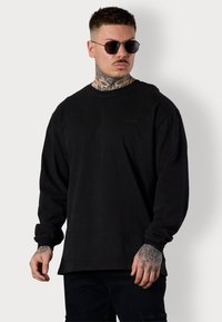 Schwarzes Langarmshirt mit geripptem Rundhalsausschnitt und dezentem Logo-Detail. Glattes Baumwollmaterial, lockere Passform, kombiniert mit schwarzen Hosen.