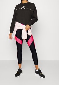 Frau, die einen schwarzen Puma-Pullover, schwarze und pinke Sportleggings, schwarze Turnschuhe und eine hellrosa Jacke, die um ihre Taille gebunden ist, trägt.