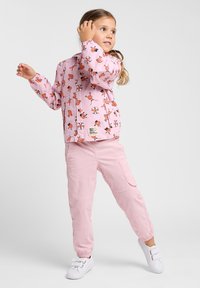 Veste rose avec des motifs de dessins animés ludiques, manches élastiques et fermeture éclair à l'avant, assortie de pantalons cargo rose clair et de chaussures blanches.