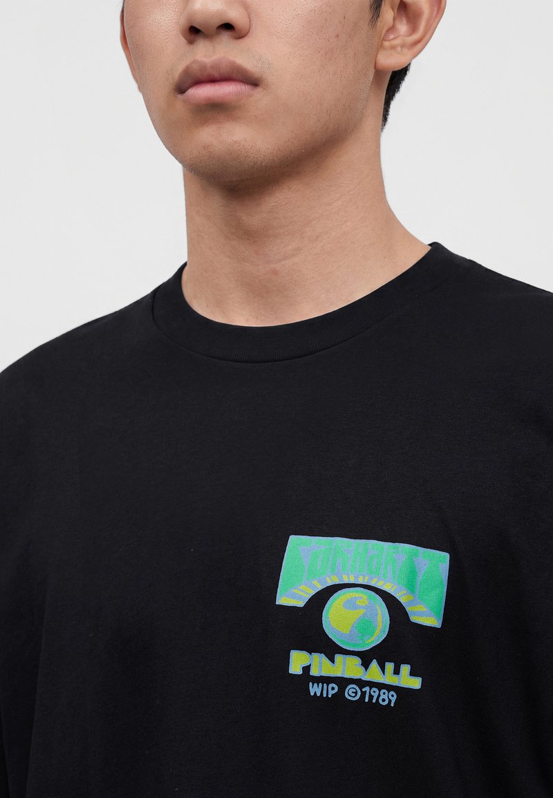 Giovane uomo che indossa una T-shirt nera con una grafica di flipper verde e blu e il testo "Pinball WIP © 1989" sul petto.