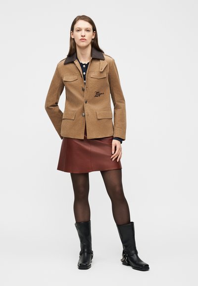 Γυναίκα που φοράει μία καφέ μπεζ ζακέτα με κουμπιά και σκούρο γιακά, μπουργκουνδί δερμάτινη φούστα, μαύρες καλσόν και μαύρες μπότες μέχρι τη μέση της γάμπας, στέκεται μπροστά σε λευκό φόντο.